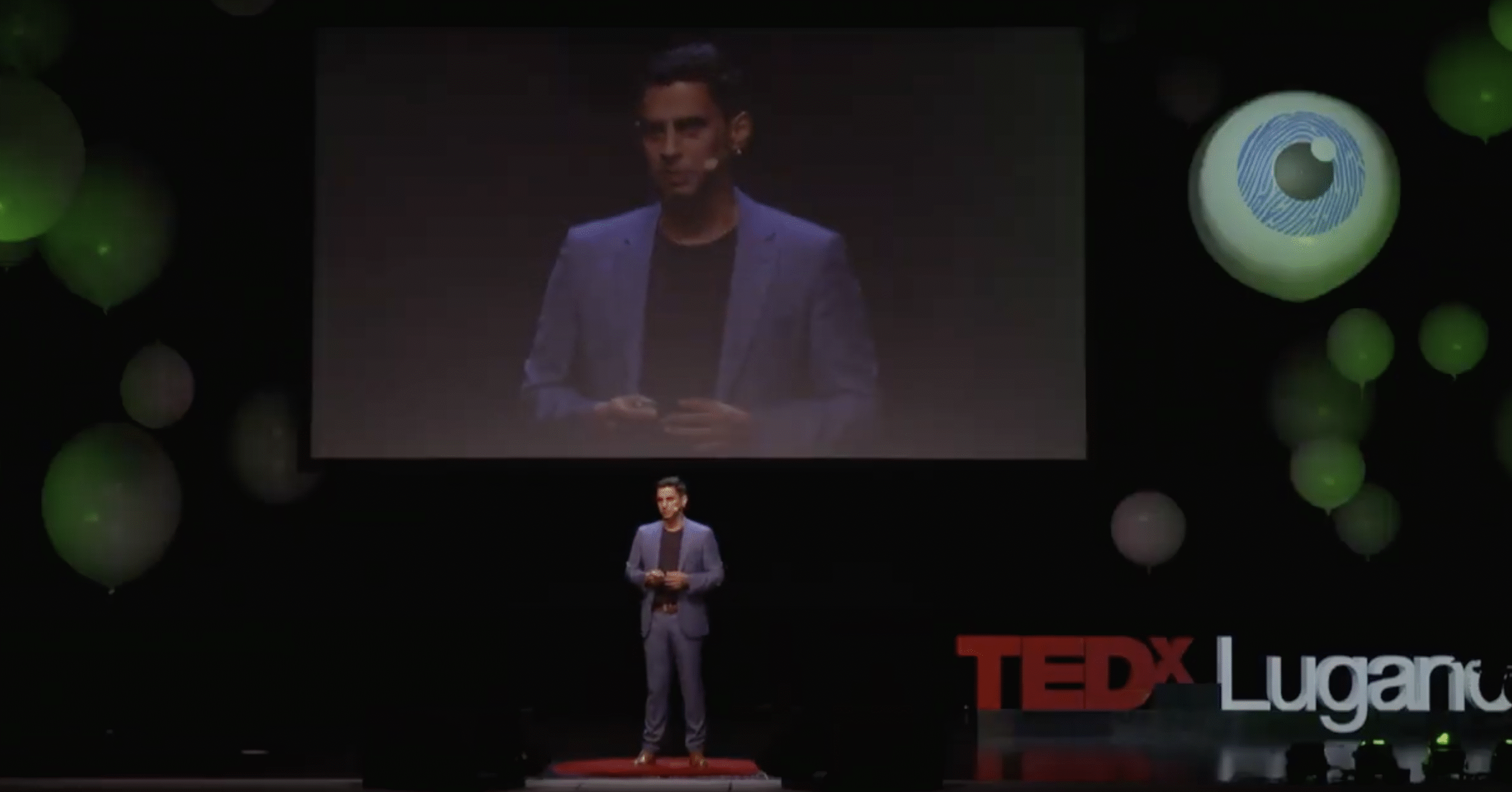 An image of Manu Kapur wearing a blue suit and presenting on stage at a Ted Talk.